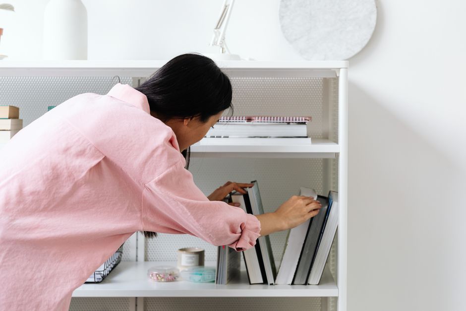 A woman in a pink shirt organizing books on a white bookshelf in an office setting.